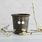 small hammered silver disc earrings hand stamped with a sun ray design on handmade french hook ear wires. The earrings are hanging on a vintage zinc bakery pot with spring blossom in front