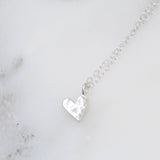 A small sterling silver, hand-pierced heart-shaped sterling silver pendant with a hammered texture on one side, displayed on a delicate chain against a marble background.